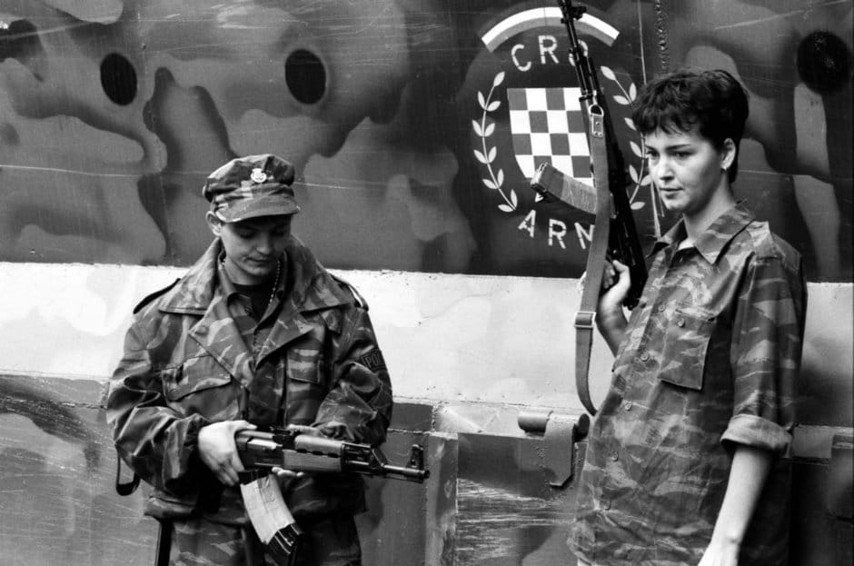 Deux femmes soldats croates au Front Gospic. Ils portent des vêtements de camouflage et des fusils ; la femme de gauche vérifie son arme et la femme de droite regarde le sol. Photographie de Robert Belosevic, septembre 1991. Image fournie par le Image of War Photography Museum.