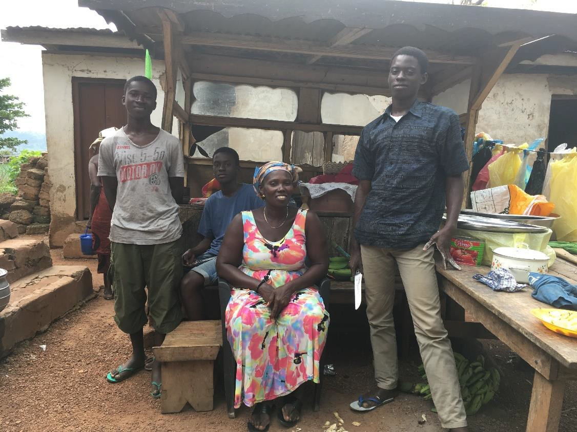  A Ghanaian woman in a bright flowery dress sits in an outdoor kitchen with her three sons around her; they range from teenaged to young adult. They are pleased with each other. Photo © Mihye Shin