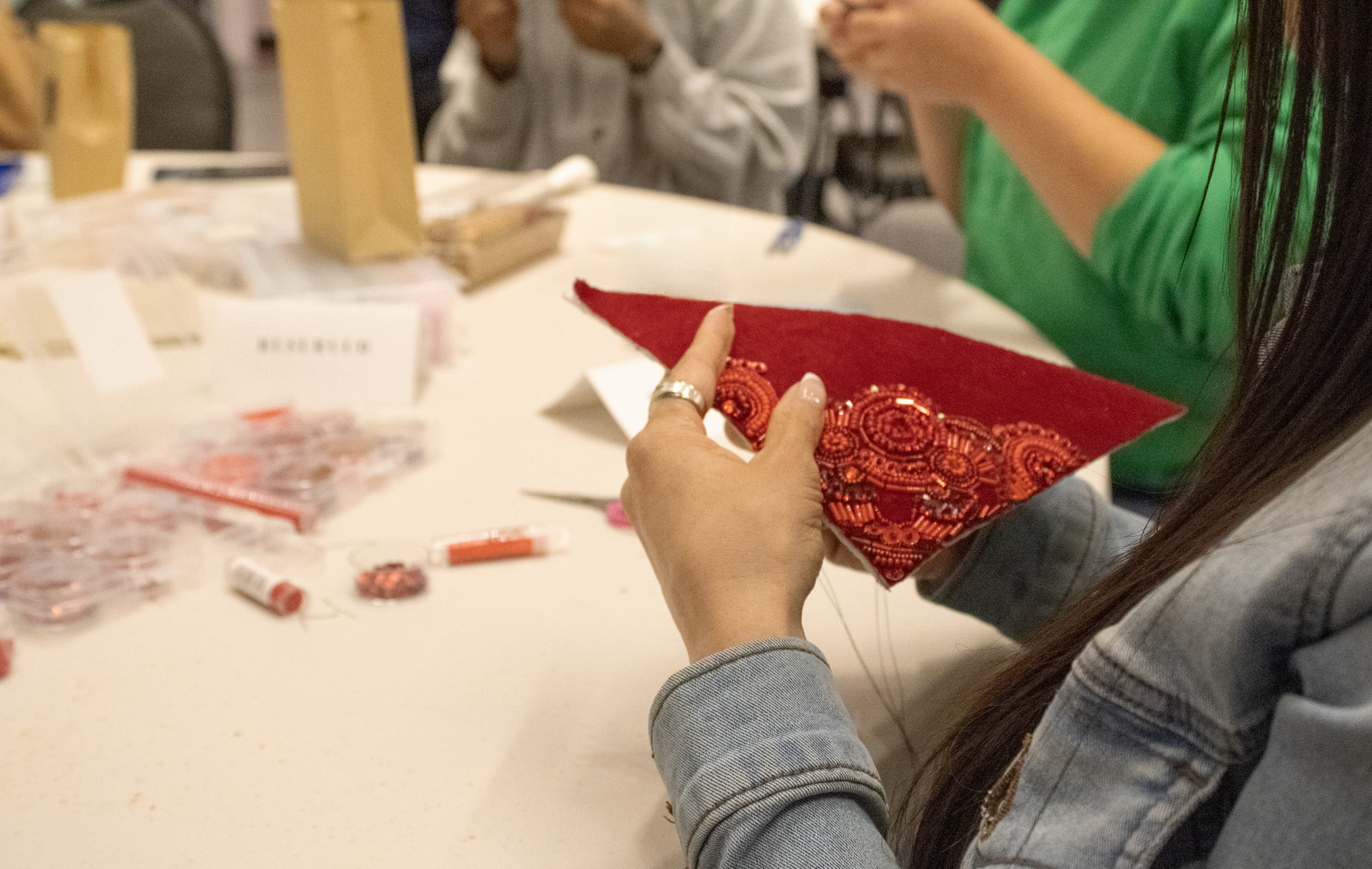 Women sit around a round table, beading; we see their hands at work. The woman closest to the camera beads orange, red, and yellow beads onto a piece of red fabric.
